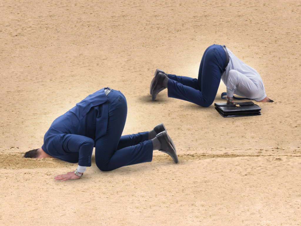 Two businessmen with their heads in the sand. One in blue suit, one in blue suit trousers and shirt and carrying black briefcase.