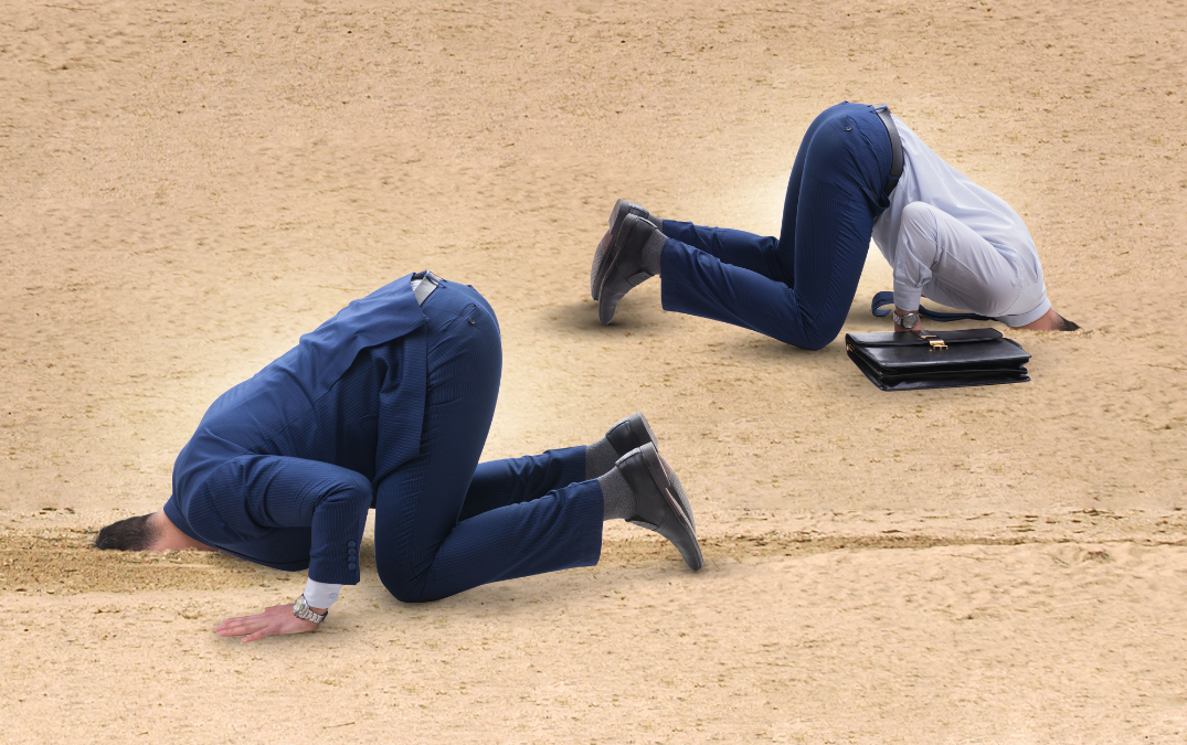 Two businessmen with their heads in the sand. One in blue suit, one in blue suit trousers and shirt and carrying black briefcase.